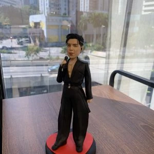 香港人氣男歌手的人像公仔，公仔身穿全黑色舞台服裝並配有入耳式耳機，手持咪高峰，站在一個紅色的心形地台上。