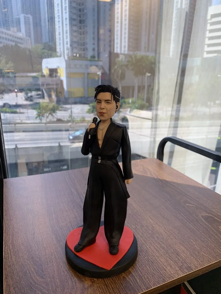 香港人氣男歌手的人像公仔，公仔身穿全黑色舞台服裝並配有入耳式耳機，手持咪高峰，站在一個紅色的心形地台上。