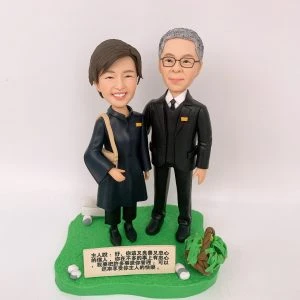 一款客製化的牧師夫婦雙人像公仔，一對男女並肩站在草地上，底座有名牌刻有聖經經文，是一份紀念他們忠心服事的榮休感謝禮物。