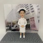 訂製醫護人像公仔，身穿白色護士制服與傳統護士帽，背景為醫院大樓與停泊的救護車，展示專業護士的典雅形象。這款公仔象徵醫生與醫護人員多年服務的感謝與榮耀，特別適合作為退休、升遷或致敬禮物。