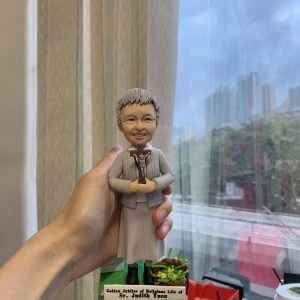 一款手持的客製化修女人像公仔，為慶祝其獻身生活50週年金慶而設，公仔手持十字架，底座刻有英文紀念字句，是送給修女或牧師等神職人員的人像公仔禮物。