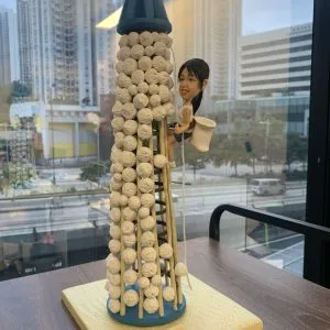 一款搶包山主題的人像公仔場景模型：中心是一座覆滿白色小包的圓柱形包山塔，頂部藍色帽狀結構，側邊設有竹梯與垂繩；一名留黑色馬尾的女性造型，著運動背心與短褲，腰間掛白色收集袋，雙手抓梯向上攀爬，表情專注帶笑。