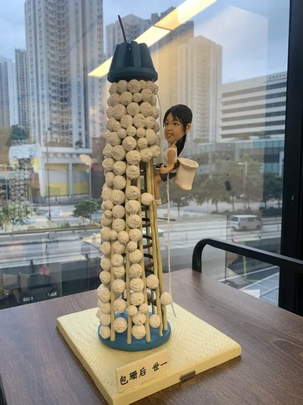 一款搶包山主題的人像公仔場景模型：中心是一座覆滿白色小包的圓柱形包山塔，頂部藍色帽狀結構，側邊設有竹梯與垂繩；一名留黑色馬尾的女性造型，著運動背心與短褲，腰間掛白色收集袋，雙手抓梯向上攀爬，表情專注帶笑。