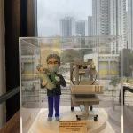 醫生人像公仔模型，身穿手術服與口罩，雙手抱著嬰兒，身旁放置新生兒保溫床與醫療設備，背景為都市高樓景色。此公仔為客製化醫生禮物，適合作為婦產科醫生、新生兒科醫護的感謝紀念品。