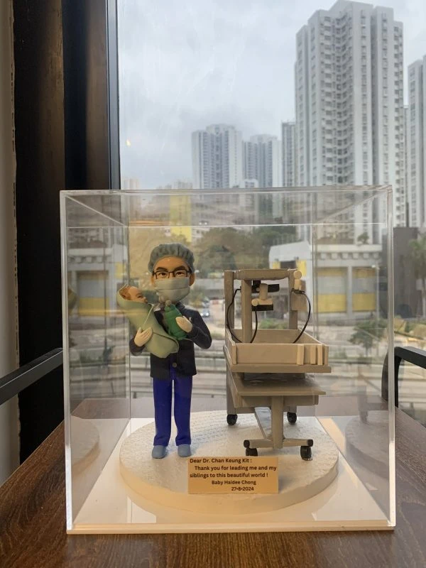 醫生人像公仔模型，身穿手術服與口罩，雙手抱著嬰兒，身旁放置新生兒保溫床與醫療設備，背景為都市高樓景色。此公仔為客製化醫生禮物，適合作為婦產科醫生、新生兒科醫護的感謝紀念品。