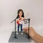 一位女性結他手的人像公仔，她身穿便服，手持紅色電結他，站在咪高峰及譜架前，地台為石板路設計。