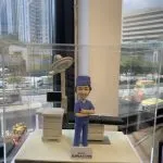 外科醫生人像公仔，身穿藍色手術服與手術帽，雙手交叉微笑站立於手術室場景。背景配有手術床、無影燈與醫療監測設備，細緻呈現專業醫護環境。此公仔為醫生、醫護人員專屬客製化禮物，適合升遷賀禮、退休紀念或醫護感謝之用。