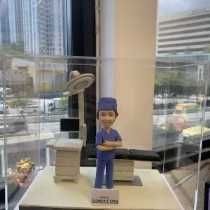 外科醫生人像公仔,身穿藍色手術服與手術帽,雙手交叉微笑站立於手術室場景。背景配有手術床、無影燈與醫療監測設備,細緻呈現專業醫護環境。此公仔為醫生、醫護人員專屬客製化禮物,適合升遷賀禮、退休紀念或醫護感謝之用。