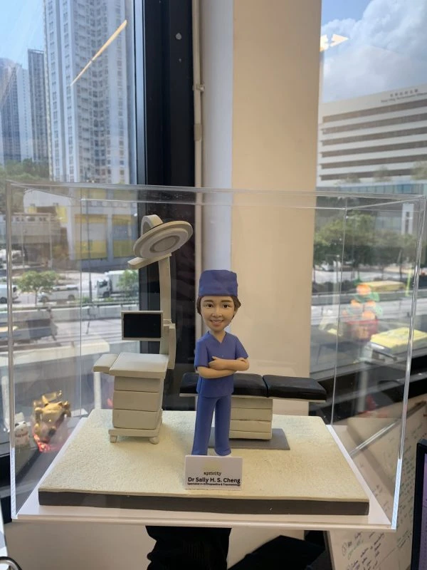 外科醫生人像公仔，身穿藍色手術服與手術帽，雙手交叉微笑站立於手術室場景。背景配有手術床、無影燈與醫療監測設備，細緻呈現專業醫護環境。此公仔為醫生、醫護人員專屬客製化禮物，適合升遷賀禮、退休紀念或醫護感謝之用。