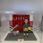 一款為高級消防隊長林龍坤訂製的退休消防人像公仔，公仔身穿制服，與一輛升降台消防車模型一同站立在油塘消防局紅色鐵閘背景前，底座刻有油塘消防局C隊的榮休祝福。