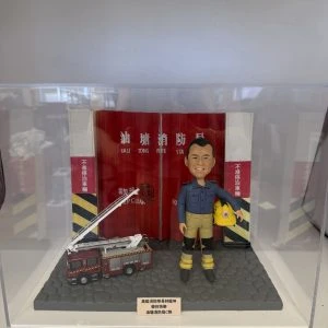 一款為高級消防隊長林龍坤訂製的退休消防人像公仔，公仔身穿制服，與一輛升降台消防車模型一同站立在油塘消防局紅色鐵閘背景前，底座刻有油塘消防局C隊的榮休祝福。