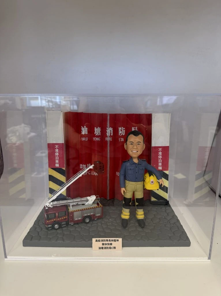 一款為高級消防隊長林龍坤訂製的退休消防人像公仔，公仔身穿制服，與一輛升降台消防車模型一同站立在油塘消防局紅色鐵閘背景前，底座刻有油塘消防局C隊的榮休祝福。