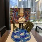 一款廚師人像公仔站在地球模型上，象徵環遊世界，是送給廚師的完美退休禮物，公仔旁有巴黎地標，背景為香港霓虹燈。