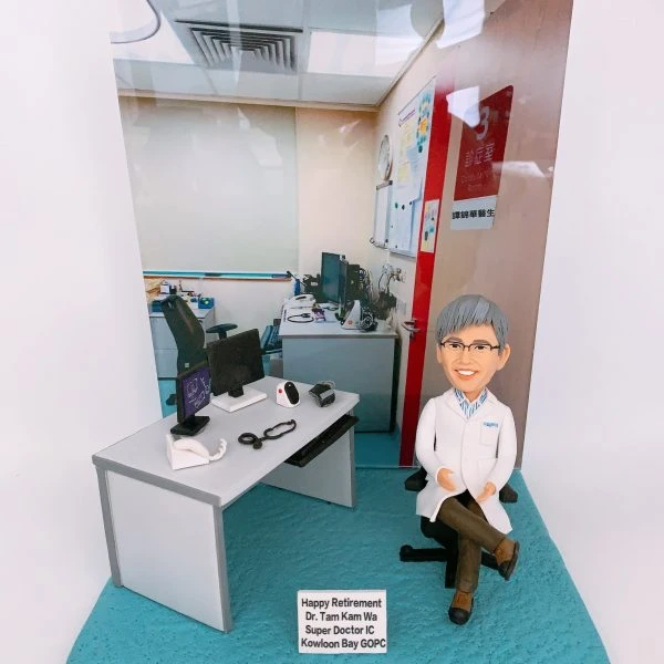 一個詳細的人像公仔，模仿了醫生譚錦華博士，坐在他的辦公室椅子上。背景展示了一間醫療辦公室，有醫療設備如電腦、聽診器及其他診斷設備。公仔穿著白色醫生袍，微笑著，保持輕鬆的姿勢。
