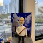 酒店主廚主題人像公仔：光頭留灰白山羊鬍的男廚師穿白色雙排釦主廚外套、黑長褲與黑鞋，右手握紅黑色長棒狀道具微笑站立。公仔置於灰色石磚紋底座，背板印有藍天白雲與高樓大廈的城市酒店天際線。背景為辦公室窗邊與高樓景色，整體具有都會質感與紀念氛圍，適合作為酒店廚師退休禮物。