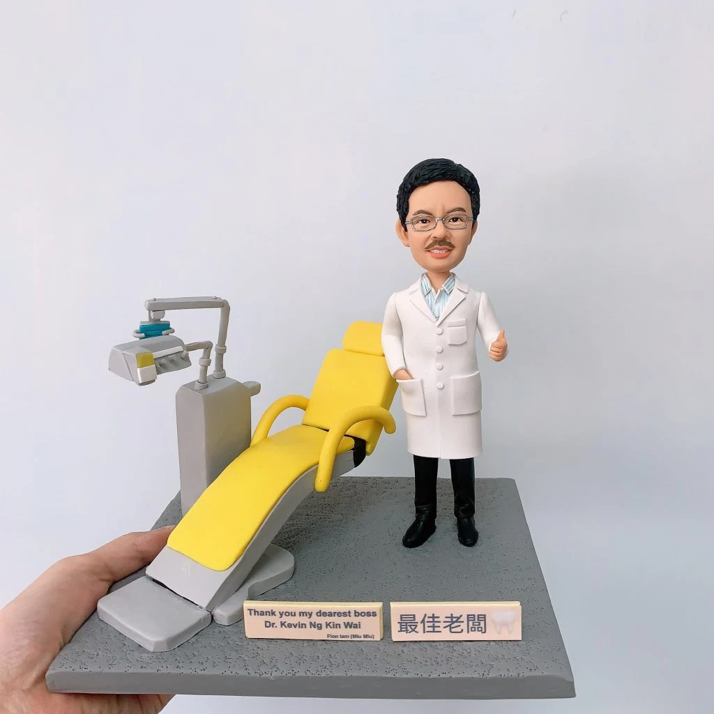 牙醫人像公仔，身穿白袍的男醫生站在黃色牙科診療椅旁，面帶笑容，右手比讚，象徵專業與親切。場景細節包含牙科專用診療椅與治療儀器，真實還原牙科診所氛圍。此專屬訂製醫生公仔適合作為牙醫禮物、醫護紀念品、退休紀念或醫療團隊感謝禮品。