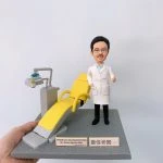 牙醫人像公仔，身穿白袍的男醫生站在黃色牙科診療椅旁，面帶笑容，右手比讚，象徵專業與親切。場景細節包含牙科專用診療椅與治療儀器，真實還原牙科診所氛圍。此專屬訂製醫生公仔適合作為牙醫禮物、醫護紀念品、退休紀念或醫療團隊感謝禮品。