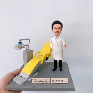 牙醫人像公仔，身穿白袍的男醫生站在黃色牙科診療椅旁，面帶笑容，右手比讚，象徵專業與親切。場景細節包含牙科專用診療椅與治療儀器，真實還原牙科診所氛圍。此專屬訂製醫生公仔適合作為牙醫禮物、醫護紀念品、退休紀念或醫療團隊感謝禮品。