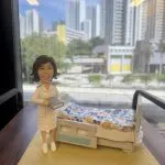 一款為護士訂製的退休禮物。精緻的人像公仔身穿白色護士服，手持記錄簿，面帶微笑地站在一張鋪有花朵圖案被子的醫院病床旁邊，是一份完美還原其工作場景、充滿敬意的紀念品。