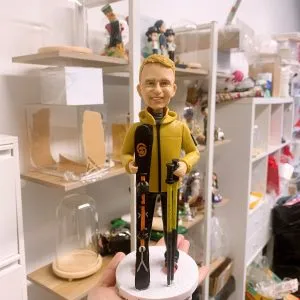 滑雪教練人像公仔，身穿黃色外套，手持滑雪杖與黑色滑雪板，站立於白色圓形底座上。精緻手工打造的人像公仔，適合作為退休禮物或紀念擺設。