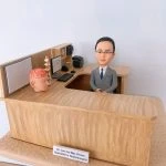 神經外科醫生人像公仔，身穿西裝的男醫生坐在診間辦公桌後方，桌面擺放大腦模型與脊椎模型，象徵專業的神經外科醫學。辦公桌上有電腦、鍵盤、醫學書籍與醫療文件，細緻還原醫生診間場景。此客製化醫護公仔適合作為醫生禮物、醫護紀念品、退休紀念或升職賀禮。