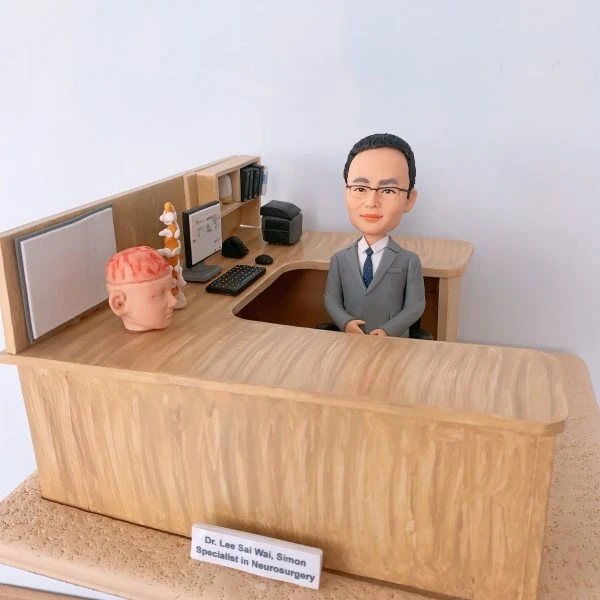 神經外科醫生人像公仔，身穿西裝的男醫生坐在診間辦公桌後方，桌面擺放大腦模型與脊椎模型，象徵專業的神經外科醫學。辦公桌上有電腦、鍵盤、醫學書籍與醫療文件，細緻還原醫生診間場景。此客製化醫護公仔適合作為醫生禮物、醫護紀念品、退休紀念或升職賀禮。