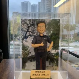 一尊警察人像公仔放置於透明展示盒內：短髮、表情親切，身穿深色短袖制服上衣與長褲，左胸口袋旁可見「POLICE」字樣；右手比讚、左手叉腰，腰間配備裝備腰帶與對講機夾。公仔站在長方形台座上，前方金色名牌印有中文姓名與「榮休之喜 享樂人生」等祝賀字樣，並標示年份區間（2019–2022）。背板為一棟帶斜屋頂與樓梯入口的建築，樹影覆蓋其上；展示盒置於木紋桌面，窗外是高樓天際線與室內燈光反射。整體氛圍為榮休致意與紀念送禮。