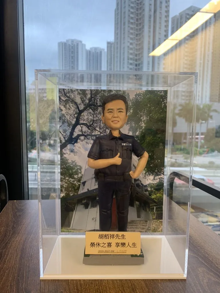一尊警察人像公仔放置於透明展示盒內：短髮、表情親切，身穿深色短袖制服上衣與長褲，左胸口袋旁可見「POLICE」字樣；右手比讚、左手叉腰，腰間配備裝備腰帶與對講機夾。公仔站在長方形台座上，前方金色名牌印有中文姓名與「榮休之喜 享樂人生」等祝賀字樣，並標示年份區間（2019–2022）。背板為一棟帶斜屋頂與樓梯入口的建築，樹影覆蓋其上；展示盒置於木紋桌面，窗外是高樓天際線與室內燈光反射。整體氛圍為榮休致意與紀念送禮。