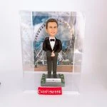 一款為外國同事訂製的人像公仔禮物，公仔是一位穿著燕尾服的男士，站立在以香港摩天輪為背景的場景中，並配有刻上祝福語的底座和透明保護盒。