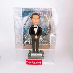 一款為外國同事訂製的人像公仔禮物，公仔是一位穿著燕尾服的男士，站立在以香港摩天輪為背景的場景中，並配有刻上祝福語的底座和透明保護盒。