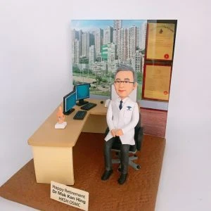 客製化醫生退休紀念人像公仔：白袍醫生坐在診症室辦公桌旁，桌上擺放雙螢幕電腦、鍵盤、滑鼠及人體模型，背景展示醫學證書與高樓大廈景觀。木質桌面與辦公椅細緻還原醫護工作環境，底座刻有「Happy Retirement Dr Mak Kan Hing HKSH OSMC」字樣，象徵醫生退休紀念與醫護專屬禮物。