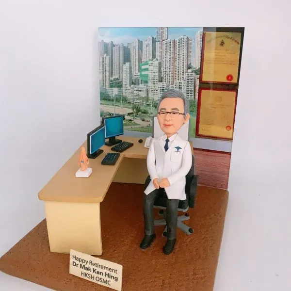 客製化醫生退休紀念人像公仔：白袍醫生坐在診症室辦公桌旁，桌上擺放雙螢幕電腦、鍵盤、滑鼠及人體模型，背景展示醫學證書與高樓大廈景觀。木質桌面與辦公椅細緻還原醫護工作環境，底座刻有「Happy Retirement Dr Mak Kan Hing HKSH OSMC」字樣，象徵醫生退休紀念與醫護專屬禮物。
