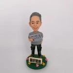 一款栩栩如生的人像公仔，呈現一位慈祥父親的造型，手中拿著帶有賭馬資訊的報紙，展現日常生活興趣。公仔底座上有「最好爸爸」字樣，並以彩色小花裝飾，傳遞濃厚的家庭溫馨氛圍與祝福。這是一份結合專屬興趣與情感的特別訂製人像公仔。