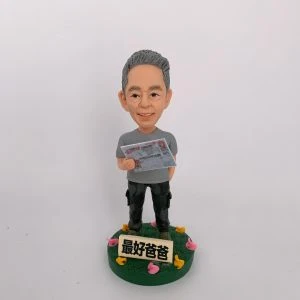 一款栩栩如生的人像公仔，呈現一位慈祥父親的造型，手中拿著帶有賭馬資訊的報紙，展現日常生活興趣。公仔底座上有「最好爸爸」字樣，並以彩色小花裝飾，傳遞濃厚的家庭溫馨氛圍與祝福。這是一份結合專屬興趣與情感的特別訂製人像公仔。