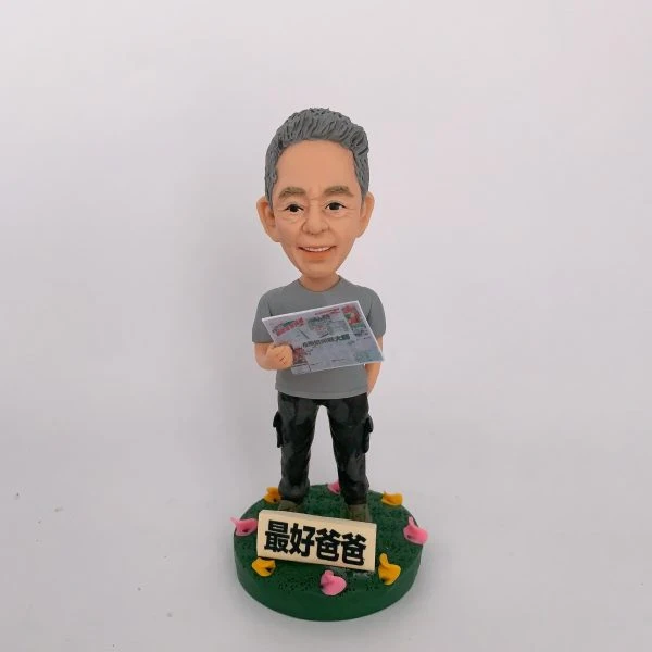 一款栩栩如生的人像公仔，呈現一位慈祥父親的造型，手中拿著帶有賭馬資訊的報紙，展現日常生活興趣。公仔底座上有「最好爸爸」字樣，並以彩色小花裝飾，傳遞濃厚的家庭溫馨氛圍與祝福。這是一份結合專屬興趣與情感的特別訂製人像公仔。