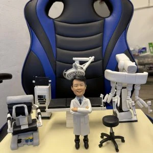 訂製醫生人像公仔:一位穿著白袍的醫生,雙手交叉站立於手術室中央,背景配備手術檯、無影燈、達文西手術機械臂(Da Vinci Surgical System)、監測設備與手術椅,完整還原微創手術場景,展現醫護專業與臨床環境。