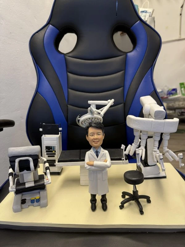 訂製醫生人像公仔：一位穿著白袍的醫生，雙手交叉站立於手術室中央，背景配備手術檯、無影燈、達文西手術機械臂（Da Vinci Surgical System）、監測設備與手術椅，完整還原微創手術場景，展現醫護專業與臨床環境。