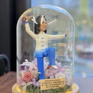 太極人像公仔展示於玻璃永生花燈罩內，公仔身穿藍白太極服，手持太極劍，單腳提腿擺出太極動作，底座裝飾有粉紫玫瑰與永生花，並附上感謝與祝福字句，作為退休禮物擺設。