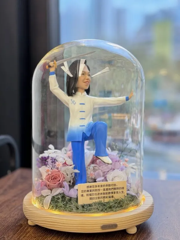 太極人像公仔展示於玻璃永生花燈罩內，公仔身穿藍白太極服，手持太極劍，單腳提腿擺出太極動作，底座裝飾有粉紫玫瑰與永生花，並附上感謝與祝福字句，作為退休禮物擺設。