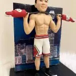 一款客製化人像公仔，公仔展現健身運動姿態，赤裸上身展現結實肌肉線條，身穿白色紅邊籃球短褲與黑色運動鞋，肩上扛著造型啞鈴，背景是香港夜景大樓燈光，象徵力量、運動與專屬紀念的結合。