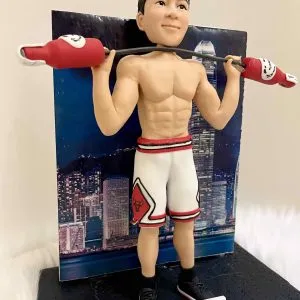一款客製化人像公仔,公仔展現健身運動姿態,赤裸上身展現結實肌肉線條,身穿白色紅邊籃球短褲與黑色運動鞋,肩上扛著造型啞鈴,背景是香港夜景大樓燈光,象徵力量、運動與專屬紀念的結合。