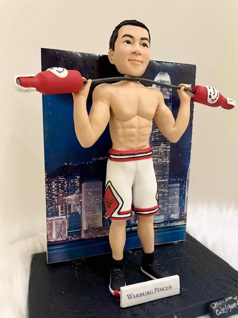 一款客製化人像公仔，公仔展現健身運動姿態，赤裸上身展現結實肌肉線條，身穿白色紅邊籃球短褲與黑色運動鞋，肩上扛著造型啞鈴，背景是香港夜景大樓燈光，象徵力量、運動與專屬紀念的結合。
