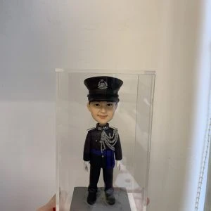 一尊穿著禮服式深色制服的警察人像公仔置於透明展示盒內，由手掌托著展示；公仔頭戴有徽章的制帽，肩章與領章清晰，胸前繫有銀灰色繩結與勳表，腰間藍色飾帶；雙手配白手套、黑色皮鞋，站在方形石磚紋台座上。背景為淺色牆面與窗邊捲簾繩。整體呈現正式典禮感，適合紀念與送禮。
