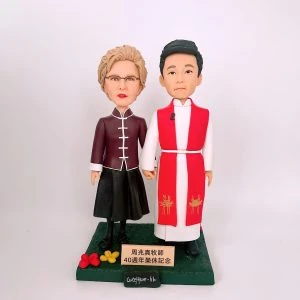 雙人牧師人像公仔並肩而立、十指相扣：右側人物著白袍配紅色聖帶與腰帶，黑鞋穩重；左側人物穿深酒紅立領中式上衣、白滾邊與黑色長裙，配戴眼鏡與短捲髮。兩人站在草綠色方形底座上，前方金色銘牌刻有「40週年榮休紀念」字樣，左前角點綴紅黃小花與品牌銘牌。整體比例優雅、細節清晰，適合作為榮休、周年與致謝的人像公仔禮物。