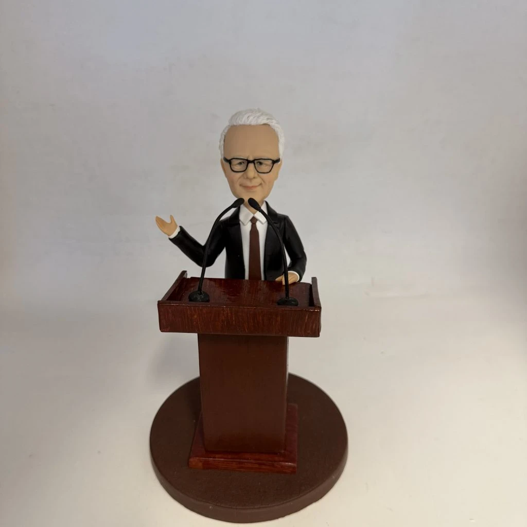 新聞發佈會主持人主題人像公仔：短白髮、戴黑框眼鏡的男士身穿黑色西裝與深色領帶，站在木紋講台後，雙手分別做講解與持講稿姿勢，面前設有兩支黑色麥克風；圓形棕色底座與素色背景突顯正式的發佈會場景。適合作為新聞發佈會主持人人像公仔的榮休禮物與展示擺設。