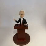 新聞發佈會主持人主題人像公仔：短白髮、戴黑框眼鏡的男士身穿黑色西裝與深色領帶，站在木紋講台後，雙手分別做講解與持講稿姿勢，面前設有兩支黑色麥克風；圓形棕色底座與素色背景突顯正式的發佈會場景。適合作為新聞發佈會主持人人像公仔的榮休禮物與展示擺設。