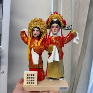 一對以戲劇角色為主題的人像公仔，身穿精緻紅色與金色華服，搭配繁複的傳統戲曲頭飾與妝容，展現舞台風華與戲劇特色。公仔細節生動，手持白布飄帶，還原戲曲動作神韻，是展現戲劇藝術與角色魅力的獨特收藏與賀禮選擇。