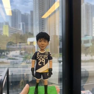 手掌上托著一尊運動主題的人像公仔，穿深藍與白色斜紋設計的球衣與短褲，胸前印有字樣「SSCPS」與號碼「29」，肩位有徽章裝飾；公仔戴黑色運動手套、穿長襪與黑色球鞋，站在亮綠色草地質感方形底座上。背景為落地窗外的城市高樓景觀與道路，室內燈光在玻璃上有反射。整體呈現青春活力與比賽氛圍。Keywords：人像公仔，運動禮物。