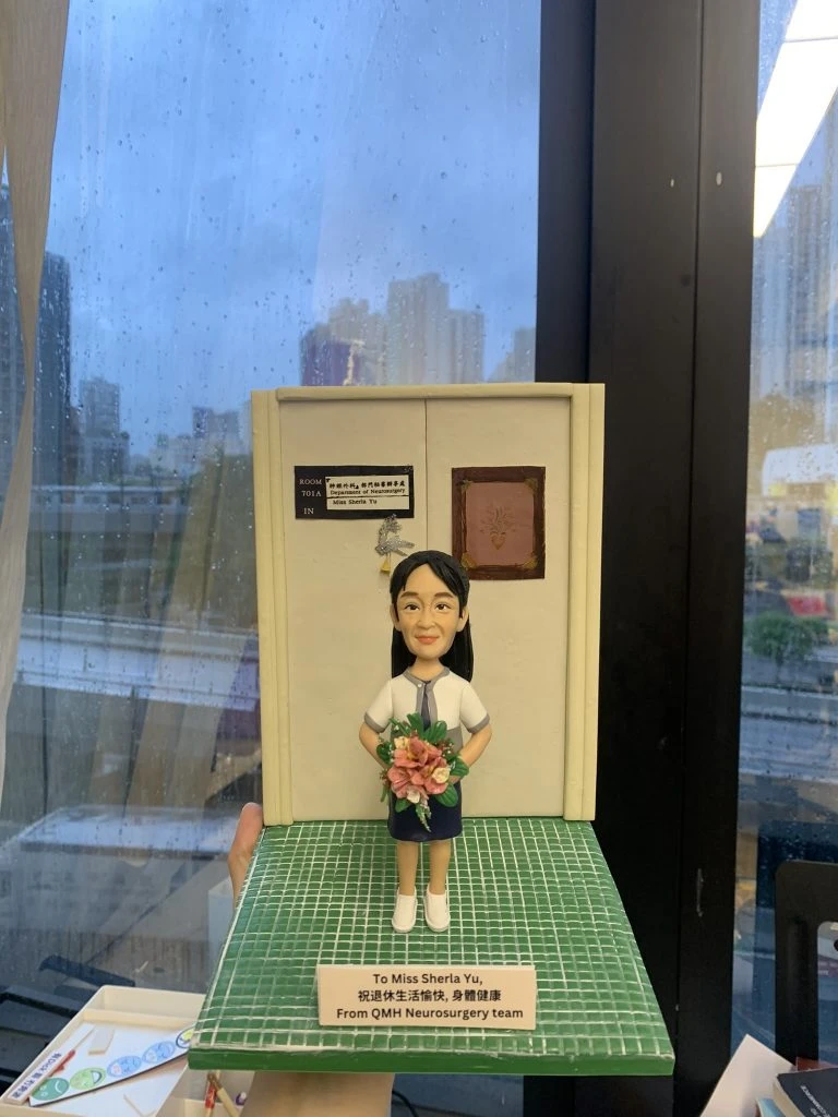 手工訂製醫護人像公仔，人物身穿醫院制服，手持花束，站立於模擬醫院場景模型前。底座刻有「To Miss Sherla Yu, From QMH Neurosurgery team」字樣，顯示來自瑪麗醫院神經外科團隊的致意。此人像公仔是專屬醫護禮物，象徵醫生與醫護人員的專業精神與感謝之情。