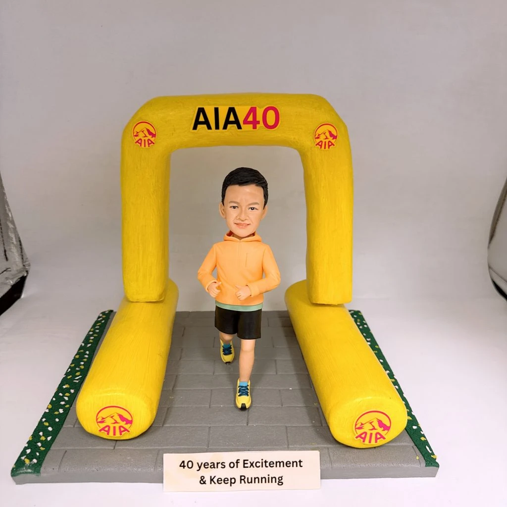 馬拉松主題人像公仔：短髮男子穿橙色連帽運動上衣、黑色短褲與黃藍跑鞋，在寫有「AIA40」的黃色充氣拱門下奔跑；拱門與兩側跑道邊緣印有 AIA 標誌，灰色路面前方放置銘牌「40 years of Excitement & Keep Running」。整體呈現AIA贊助的馬拉松比賽終點氛圍。